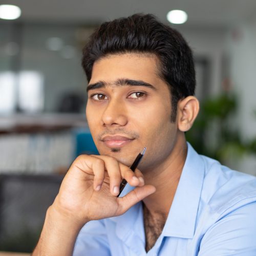 Portrait of young Indian businessman or student sitting with pen and looking at camera in office. Businessman concept